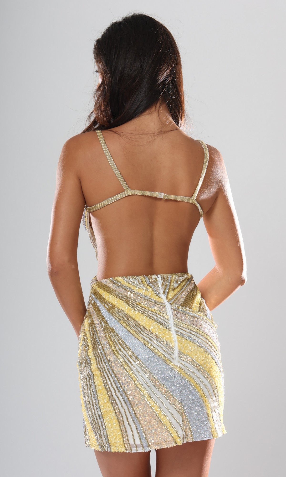 A woman with long brown hair stands facing away from the camera, wearing the Jovani 53768 short homecoming dress—a backless design embellished with gold, silver, and white sequined beads in striking stripes.